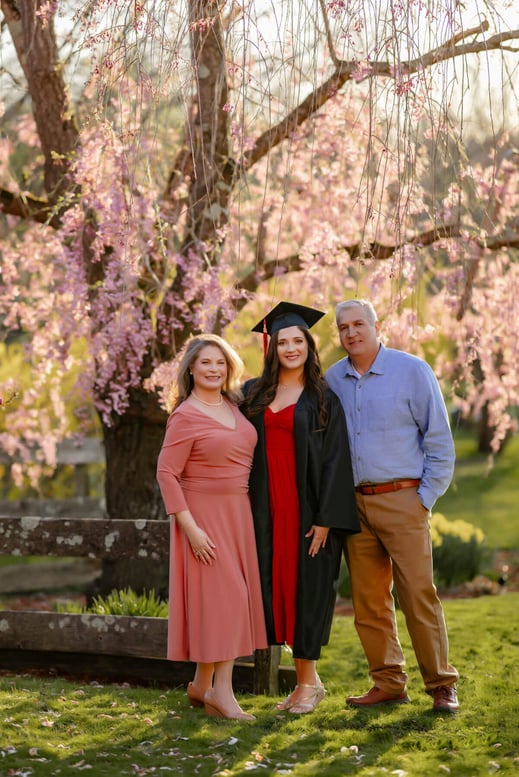 High school graduate senior girl in her cap & gown with her mom & dad with a pink flower tree as background, at Lexington, Massachusetts