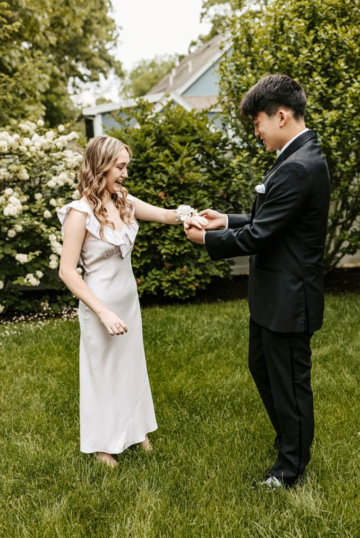 High school senior girl dressed in a grey long dress with her boyfriend wrapping a bouquet flower at her wrist, in a green backyard, at Lexington, Massachusetts
